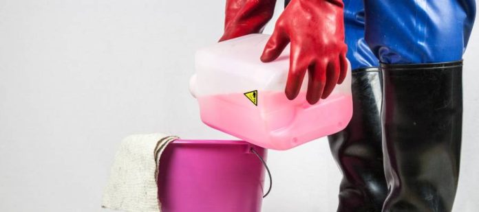 A worker wearing rubber gloves, rubber pants, and chemical boots pours a pink chemical into a bucket.