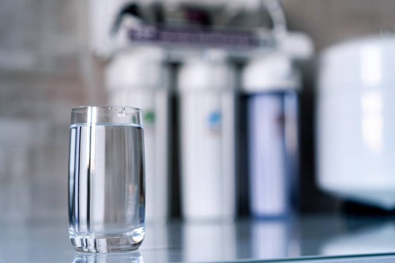 A full glass of water on a countertop with a whole home water filtration system in the blurred background.