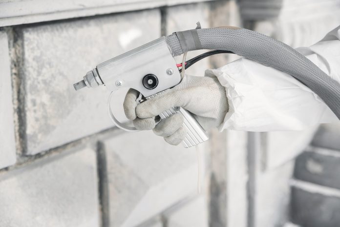 A white-gloved hand holding a sandblaster nozzle and applying a stream of abrasive media to a gray stone wall.