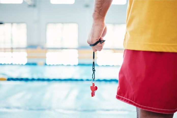 A lifeguard standing in front of a pool. Only his lower back and left arm are visible. He grips a red whistle.