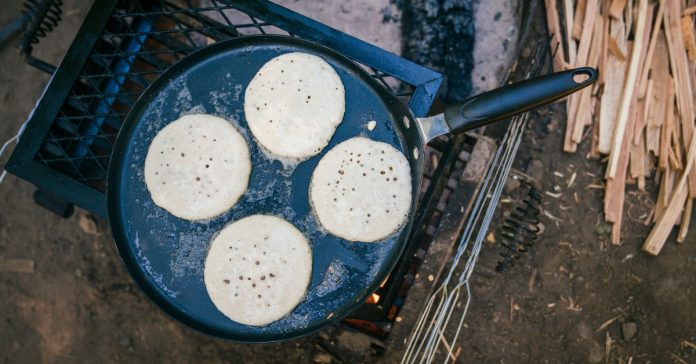 A large, greasy skillet placed on a rack over a campfire, with four pancakes bubbling and cooking over it.