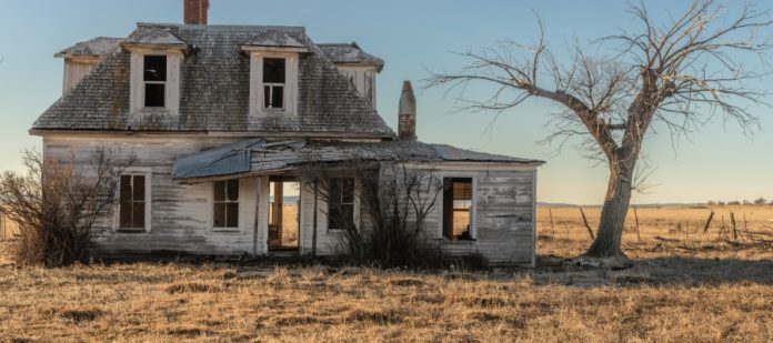 An old prairie house is beat up with no one living in it. There are also dead trees and grass surrounding it.