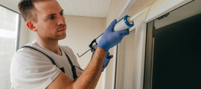 A young man stands on a ladder and uses a caulk gun to seal the gap above a sliding glass patio door.