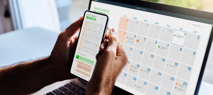 A close-up of a man holding up his smartphone with a calendar. He’s in front of his laptop with the same calendar.