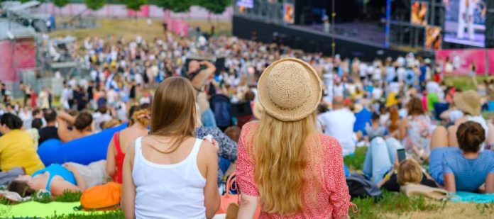 Two women are sitting on a blanket in a crowded field of people. They are listening to an outdoor music festival.