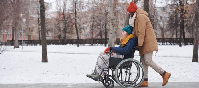 A young man in a jacket pushes a young woman in a coat in a wheelchair through the park. Snow is on the ground.