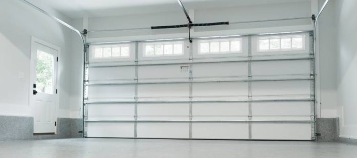 Interior view of a big, clean, and empty garage with white walls, a large white garage door and an entry door to the left.