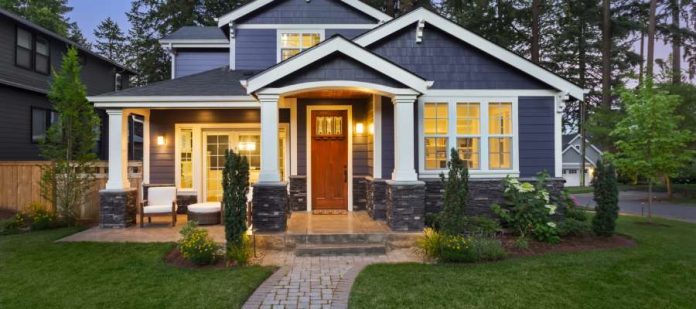 The exterior of a beautiful blue house lit up from the inside with white columns and lush greenery and landscaping.