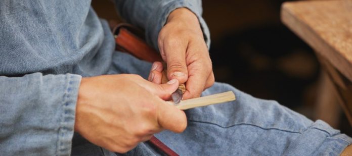 Close-up of someone cutting a thin piece of wood with a small knife. The person wears a denim shirt and apron.