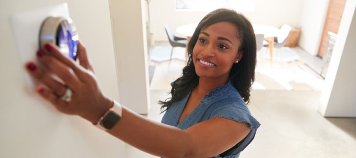 A woman in her home is happily adjusting the thermostat. Behind her is a living room painted white.