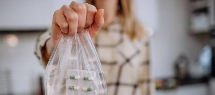 A woman's hand is seen in close-up holding a clear plastic baggie filled with expired medications, ready for disposal.