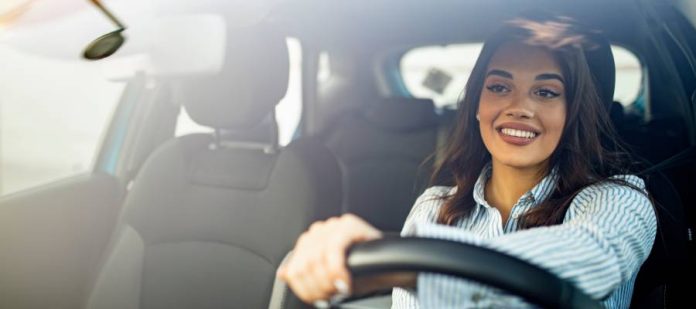 A woman wearing a striped button-down shirt drives a hatchback sedan with one hand on the wheel while smiling.