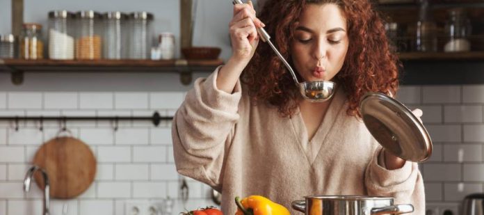 A young woman with auburn curly hair is cooking soup in a kitchen. She's blowing on the soup in a ladle to cool it down.