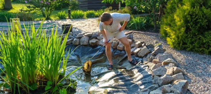 A young man in a long-sleeve shirt and shorts cleaning a backyard pond with a net. The net contains debris from the water.