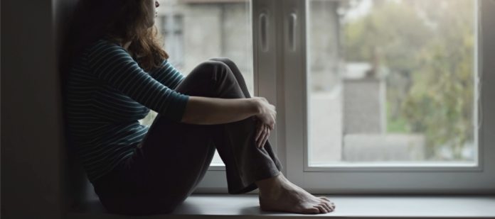 A dejected woman sits on a window ledge with her knees to her chest while looking out the window sorrowfully.