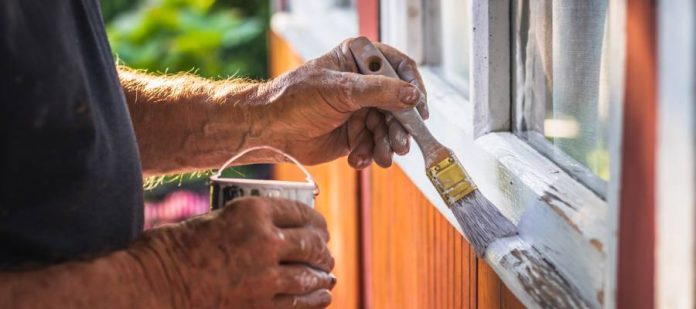 A close-up view shows a person using a skinny paint brush to paint white paint on wooden window trim.