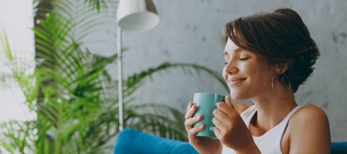 A woman holds a mug near her face and smiles with her eyes closed. She wears a white tank top and sits on a blue couch.