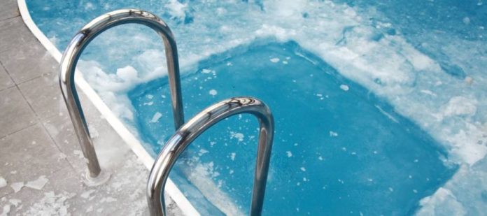 A swimming pool covered in ice and slush. An ice hole revealing blue water is near the pool's metal handrails.