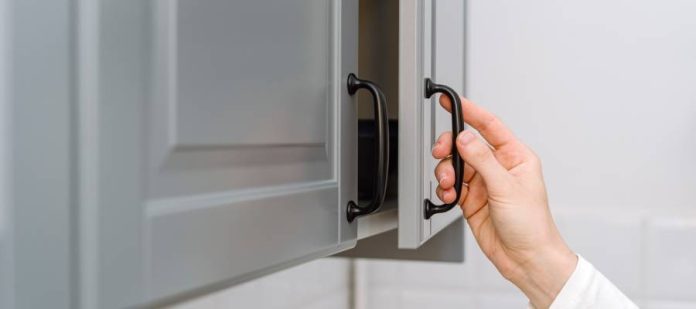 A woman's hand opening a blue-gray kitchen cabinet with a black handle. The woman wears a long sleeve white shirt.