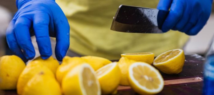 A man's hands, covered with food preparation gloves and holding a cleaver, is shown cutting many lemons in half.