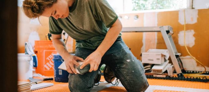 A woman in a t-shirt and jeans sands a piece of tile in an unfinished bathroom filled with renovation tools and supplies.