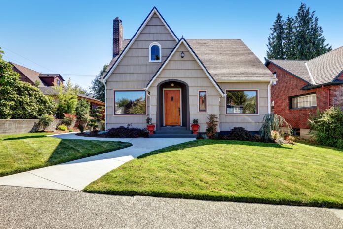 A small craftsman-style home with a well-maintained lawn and a row of hedges and trees beside it. The sky above is blue.