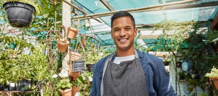 A man in a gray apron is standing in a store full of potted plants. He's holding a small paper sign that says 