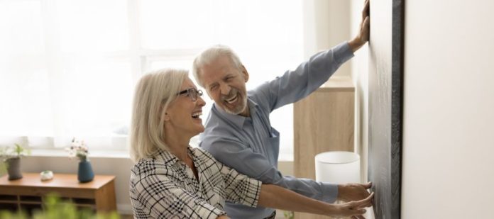 An older man and woman aligning a large frame onto a wall. They are both smiling while using two hands to set the frame.