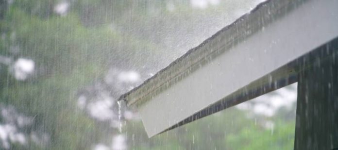 A close-up of heavy storm rain running off a house's roof. Another roof is visible in the background.