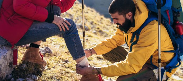 A woman sits on a rock at the top of a mountain. A man kneels in front of her and wraps her ankle in a bandage.