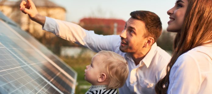 A mother, father, and toddler sit next to a large solar panel in their yard. The father points to the sky above the solar panel.