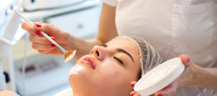 A medical spa client laying on the exam table with a licensed esthetician using a facial brush to apply a face mask.