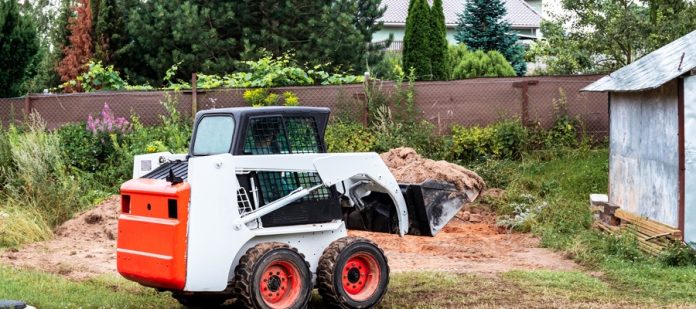 A white and orange skid steer with a bucket full of dirt drives across grass in a fenced area with trees outside of it.