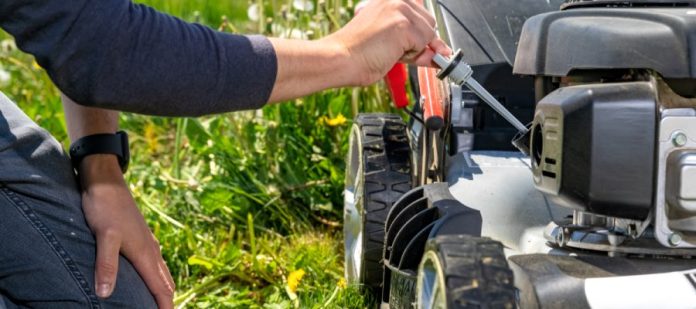 A man kneels in the grass next to a push lawn mower and checks the oil. There is tall grass behind the lawn mower.
