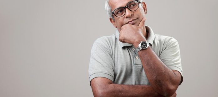 A man with gray hair rests his hand against his face. He also wears glasses, a watch, and a gray t-shirt.