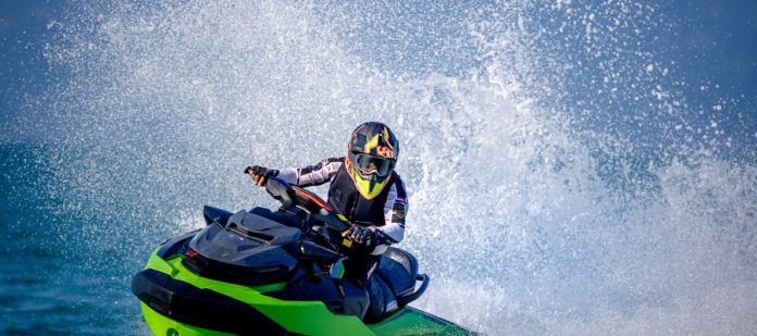 A man in a helmet, goggles, gloves, and safety vest making waves while driving a black and green jet ski.