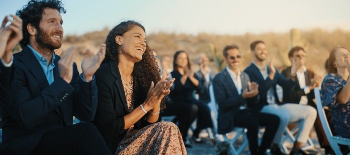 A large group of people sitting in chairs at an outdoor wedding. They are all smiling and clapping.