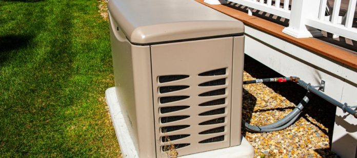A brown residential generator on a white pad sitting outside next to a deck with a wooden railing on a sunny day.