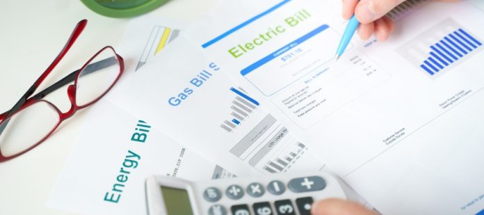 A person holds a pen in one hand and uses a calculator with the other. Energy, gas, and electric bills sit on the table.