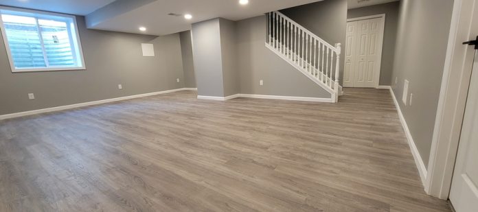 An empty, newly-finished basement of a modern home, including vinyl plank flooring, grey walls, and a white staircase.