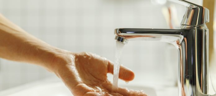 A man places one hand under a chrome faucet that's turned on. Water runs off his fingers and into the sink.