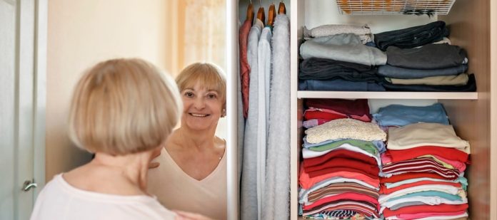 A woman smiles into her closet's sliding mirror door. Clothing stacks and a hanger compartment sit behind the door.