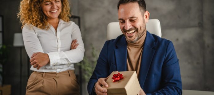 A man in a blue blazer smiles as he opens a cardboard gift box with a red bow on top in front of his colleague.