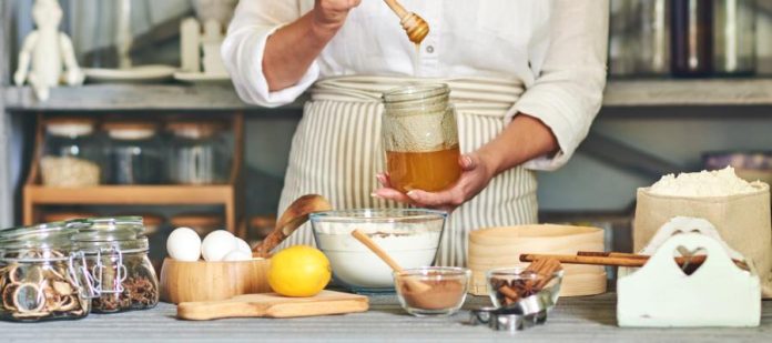 A close-up of a woman holding a jar of honey and preparing dough with an array of ingredients for baking in the kitchen.