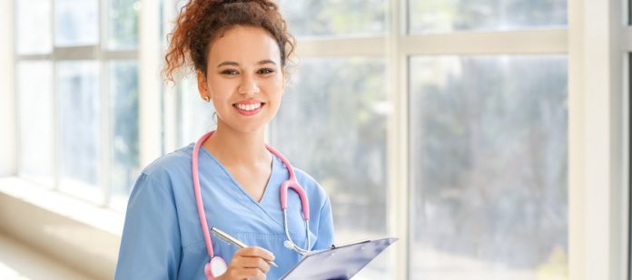 A young woman in blue scrubs with a pink stethoscope around her neck holds a pen and a blue clipboard.