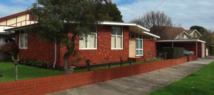 A red brick mid-century house sits behind pruned rose bushes, with large windows and a carport on the right side.