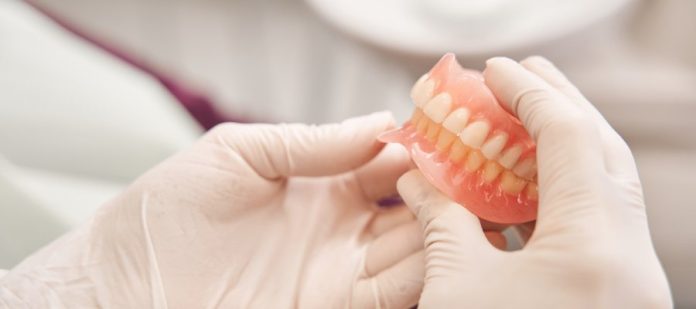 A dentist, wearing white gloves, holds a set of brand-new dentures under a light in the dentist's office.