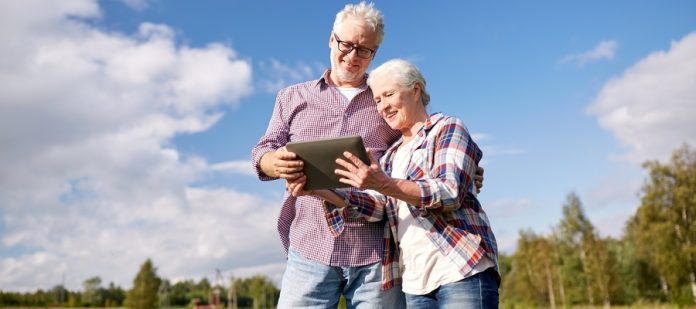 A happy senior couple leaning against each other and looking at a tablet in a large open field under a blue sky.