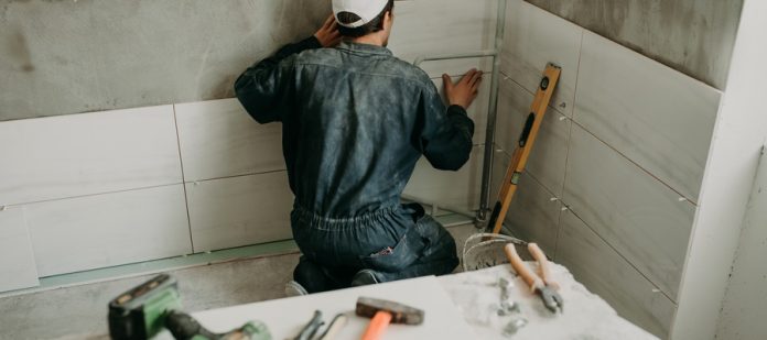 A contractor installing a tile wall in a bathroom. A pile of tools is on the counter behind the contractor.