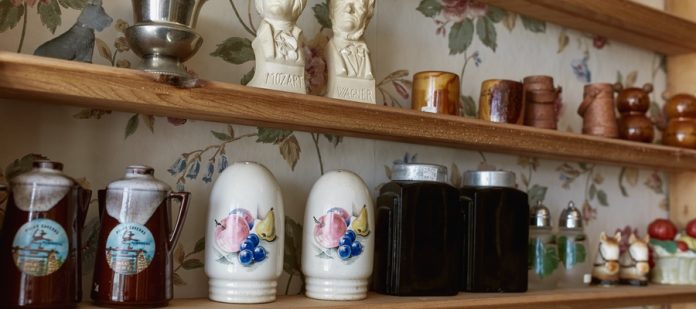 Several salt and pepper shakers on a wooden shelf. Some shakers look like historical figures, kettles, and other items.